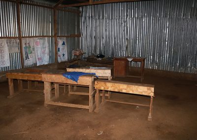 Desks on a dirt floor with metal walls which is a kindergarten class for Our Lady of Grace School in Kenya