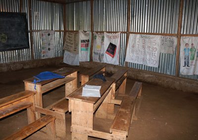 Desks on a dirt floor with metal walls which is a kindergarten class for Our Lady of Grace School in Kenya